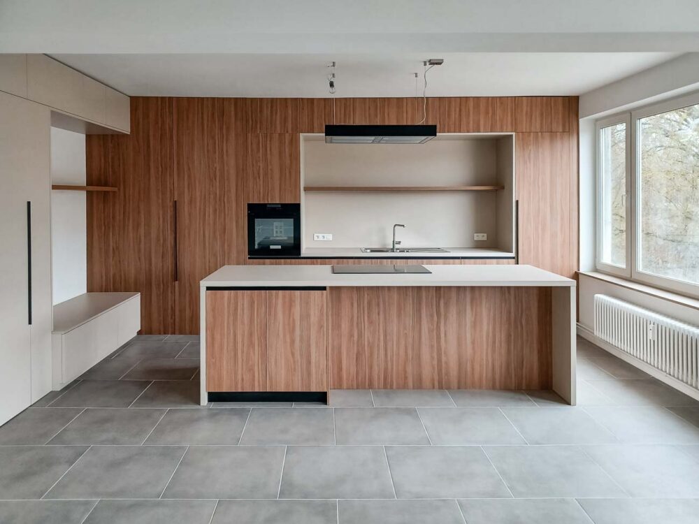 Cuisine en bois et gris beige avec long mur entièrement meublé de rangements sur mesure avec portes invisibles donnant accès aux autres pièces