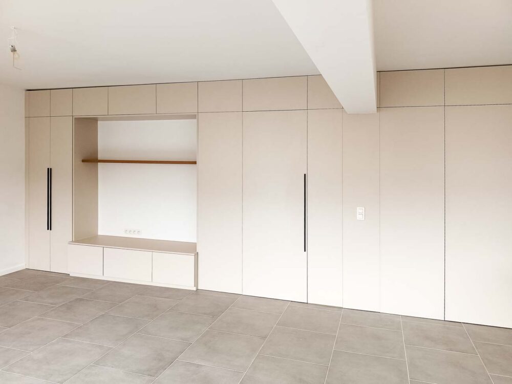 Meubles de living beiges entièrement adaptés à la pièce, avec vestiaire et portes invisibles donnant accès à d’autres pièces