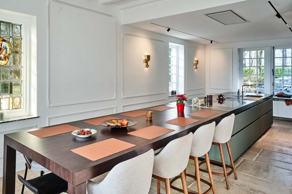 Cuisine spacieuse haut de gamme avec plan de travail en pierre naturelle vert foncé quartzite, îlot central en verre vert olive, détails en cuivre (Inox PVD) et composition murale en noyer foncé