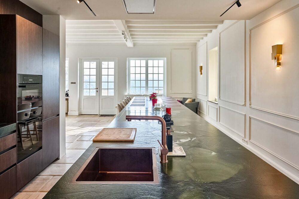 Cuisine spacieuse haut de gamme avec plan de travail en pierre naturelle vert foncé quartzite, îlot central en verre vert olive, détails en cuivre (Inox PVD) et composition murale en noyer foncé