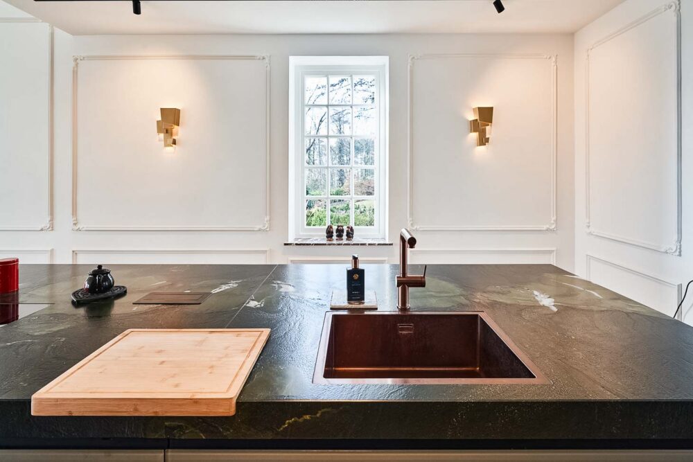 Cuisine spacieuse haut de gamme avec plan de travail en pierre naturelle vert foncé quartzite, îlot central en verre vert olive, détails en cuivre (Inox PVD) et composition murale en noyer foncé