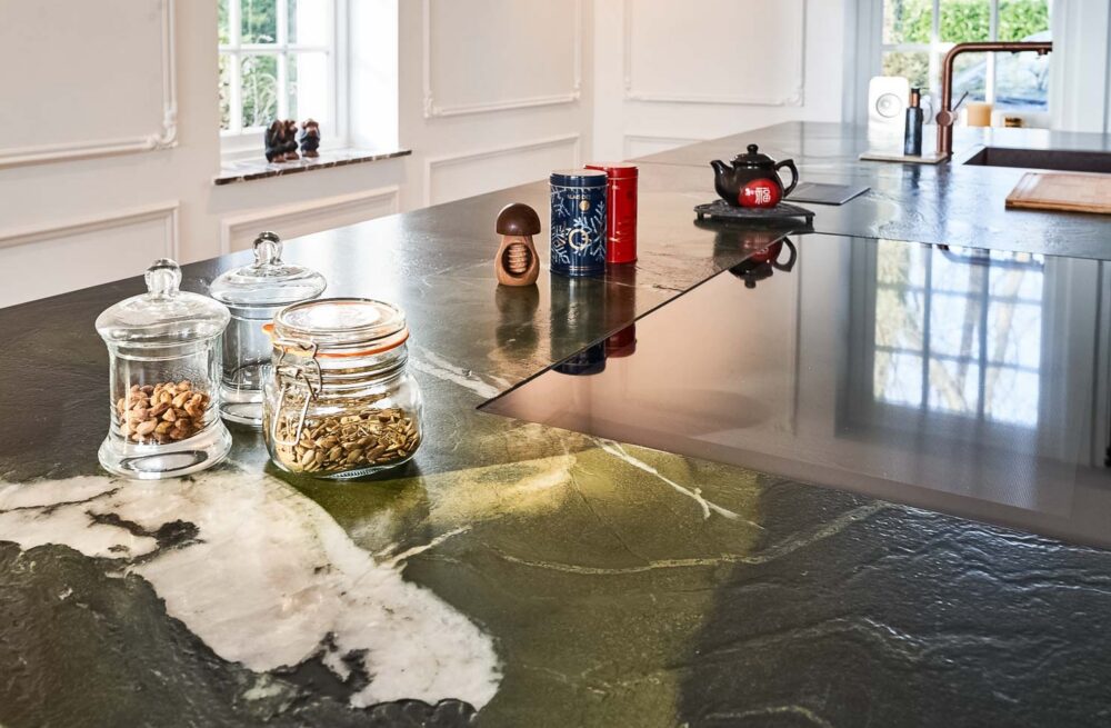 Cuisine spacieuse haut de gamme avec plan de travail en pierre naturelle vert foncé quartzite, îlot central en verre vert olive, détails en cuivre (Inox PVD) et composition murale en noyer foncé