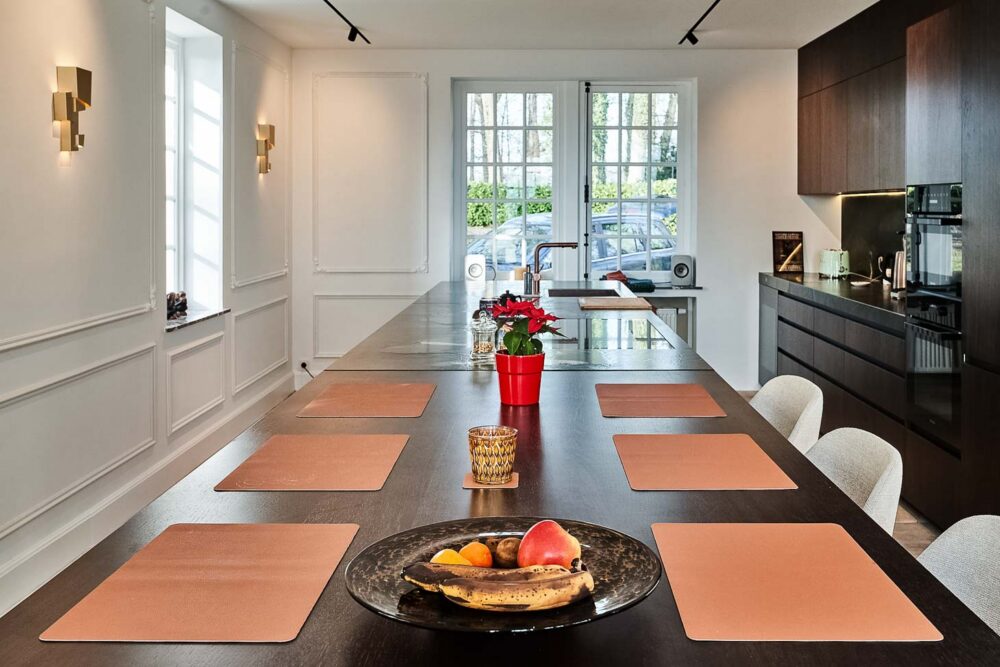 Cuisine spacieuse haut de gamme avec plan de travail en pierre naturelle vert foncé quartzite, îlot central en verre vert olive, détails en cuivre (Inox PVD) et composition murale en noyer foncé