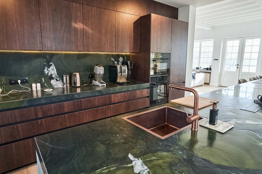 Cuisine spacieuse haut de gamme avec plan de travail en pierre naturelle vert foncé quartzite, îlot central en verre vert olive, détails en cuivre (Inox PVD) et composition murale en noyer foncé