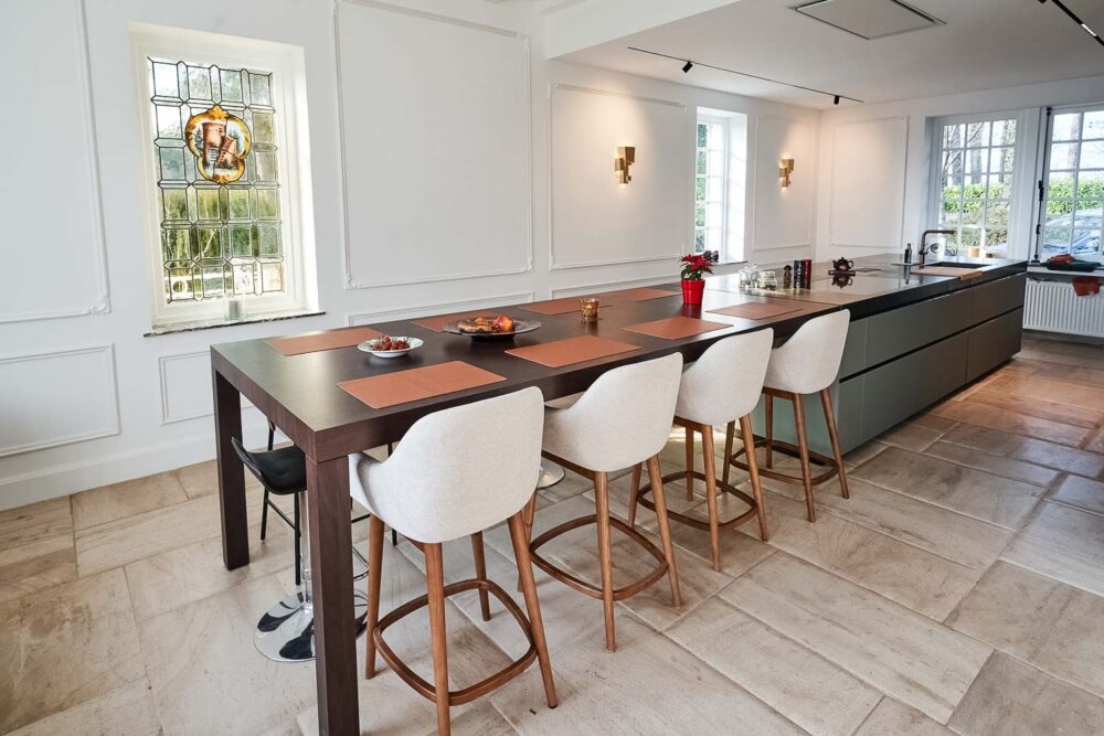 Cuisine spacieuse haut de gamme avec plan de travail en pierre naturelle vert foncé quartzite, îlot central en verre vert olive, détails en cuivre (Inox PVD) et composition murale en noyer foncé