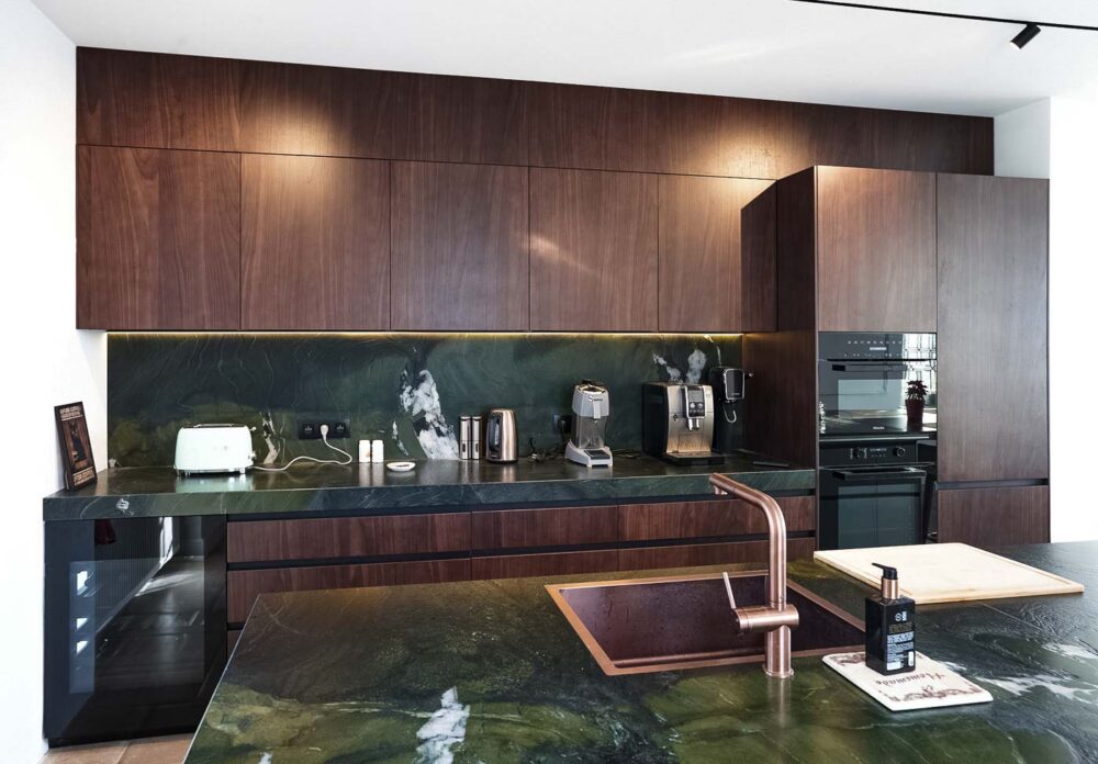 Cuisine spacieuse haut de gamme avec plan de travail en pierre naturelle vert foncé quartzite, îlot central en verre vert olive, détails en cuivre (Inox PVD) et composition murale en noyer foncé
